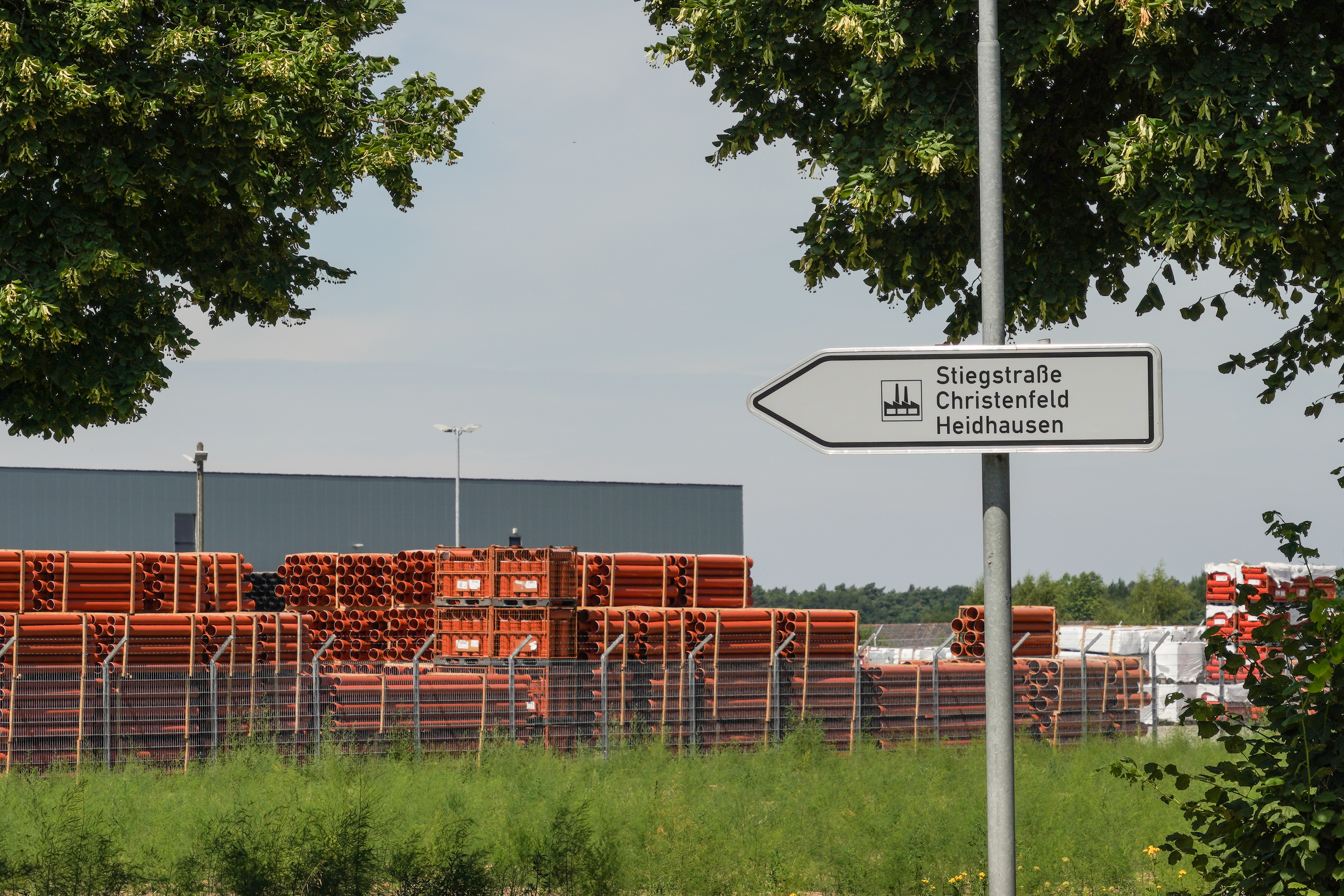 Weißes Schild an Metallstange mit Fabriksymbol in schwarz, weiß und Schrift rechts davon untereinander " Stiegstraße", "Christenfeld", "Heidhausen"", mit gestapelten, roten Rohren hinter Zaun vor grauer Halle im Hintergrund, vor Zaun grüner Pflanzenbewuchs, am Bildrand dichter, grüner Bewuchs von Baumkronen